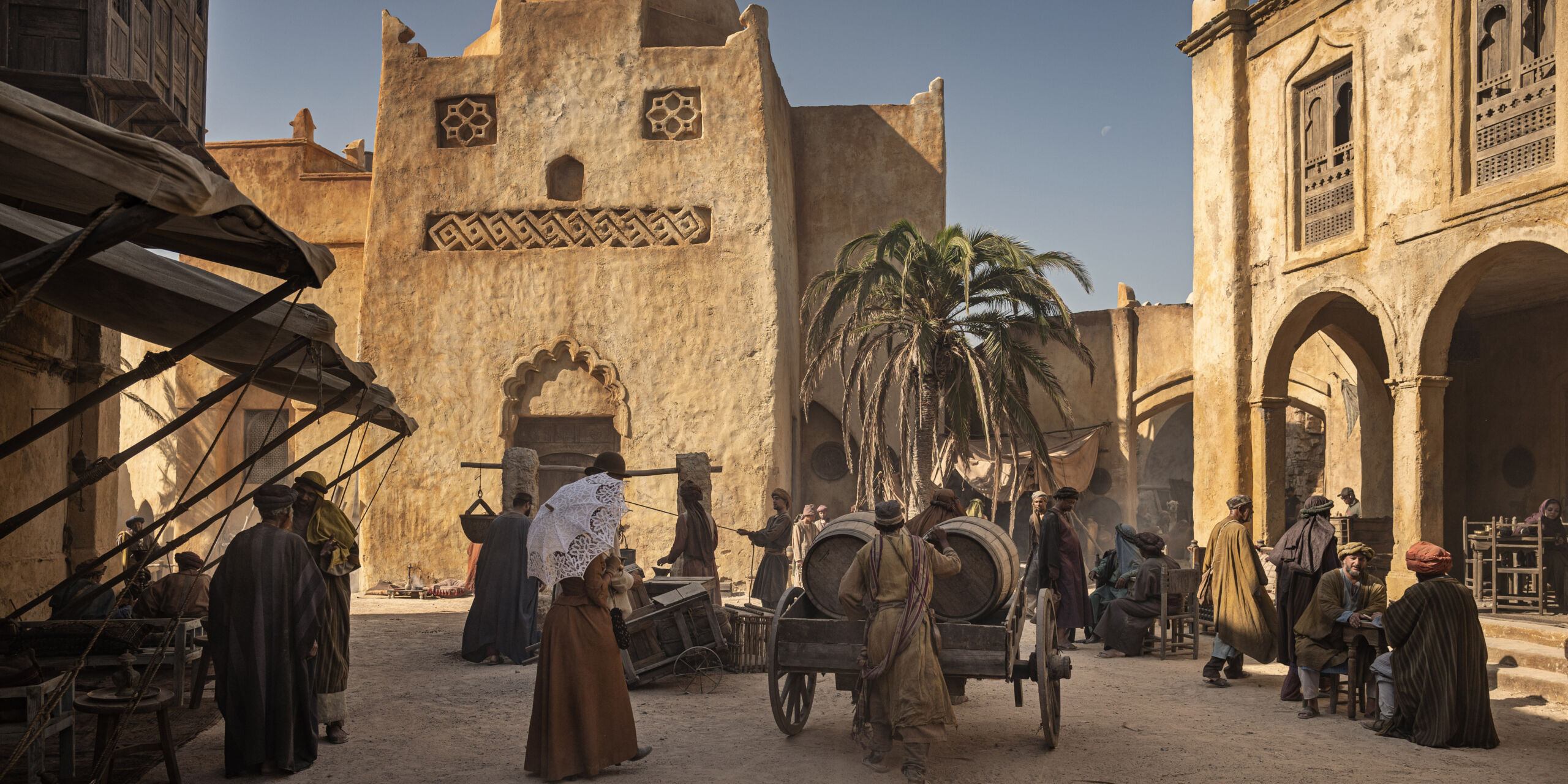 Television with adobe structures, possibly in North Africa, and men wearing turbans and dresses.  A woman walks with a white parasol.