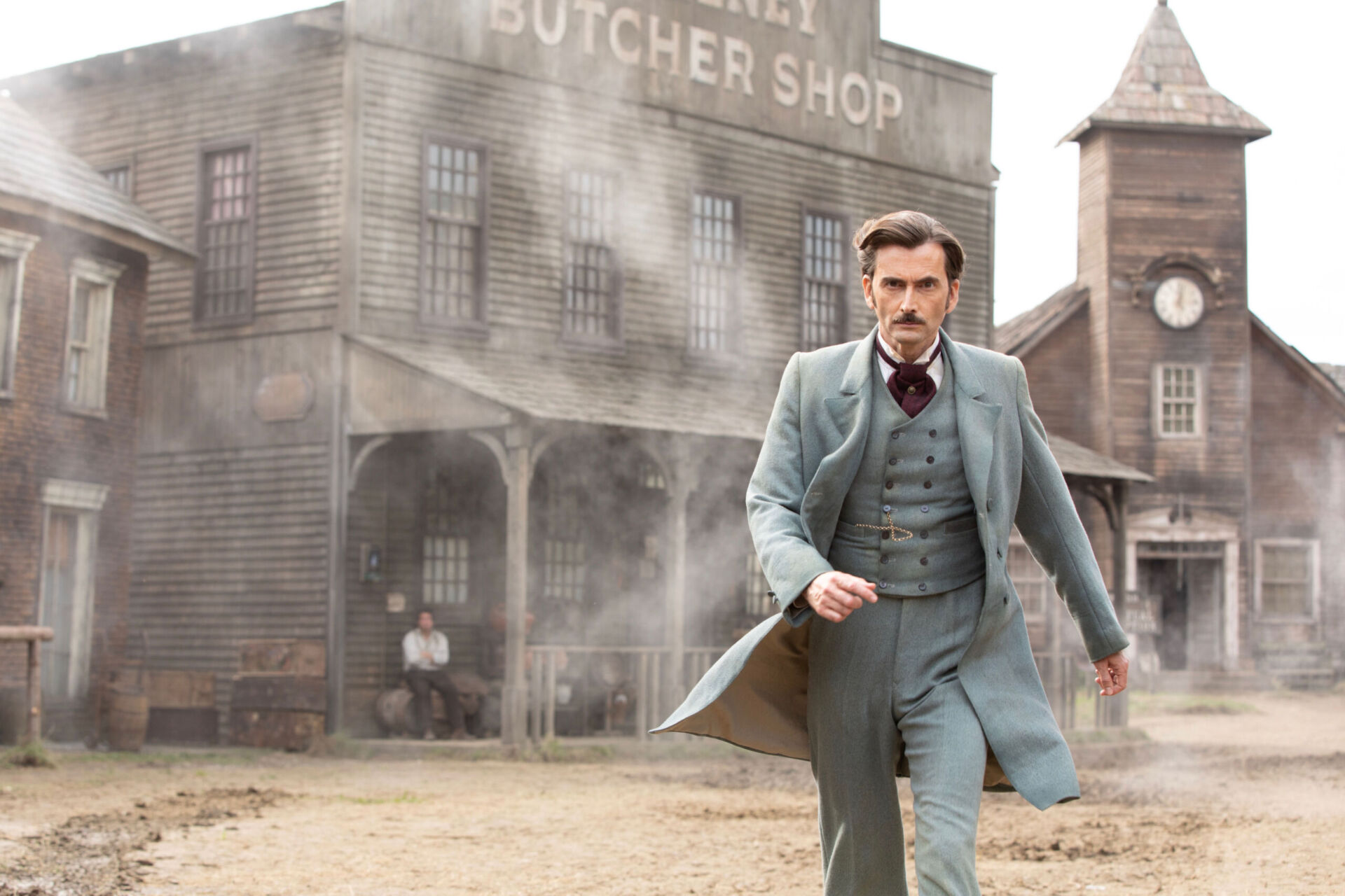 A man in a 19th-century three-piece suit walks through a dusty plaza with American wooden structures, including a butcher's shop and a church or town hall